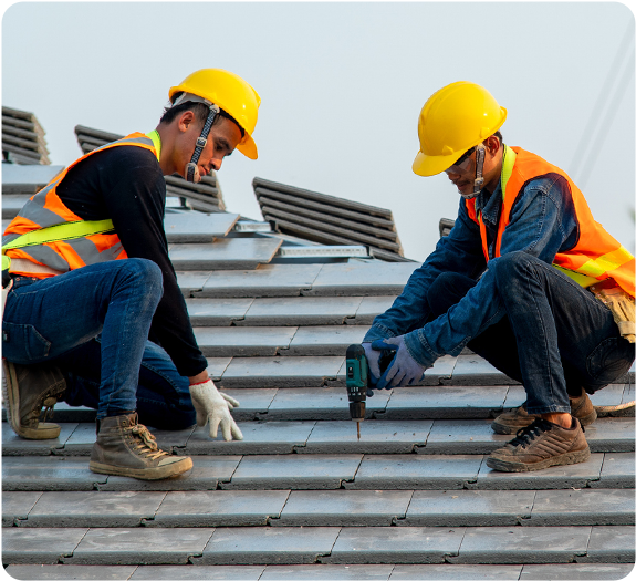 Two male roofers wearing yellow hard hats and safety vests drilling new shingles into a roof