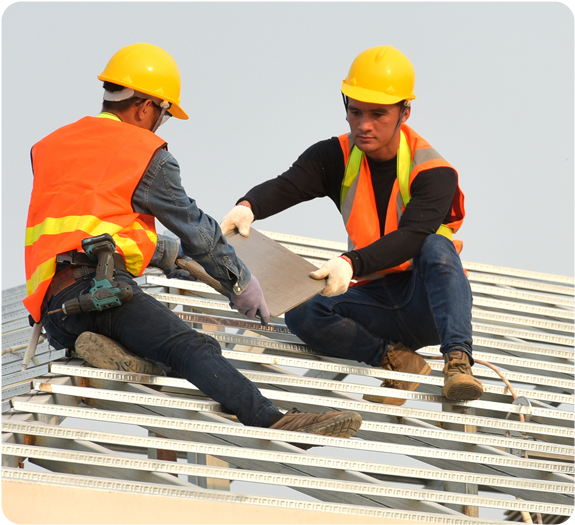 Two male roofers wearing safety gear holding a shingle on the roof as they install it