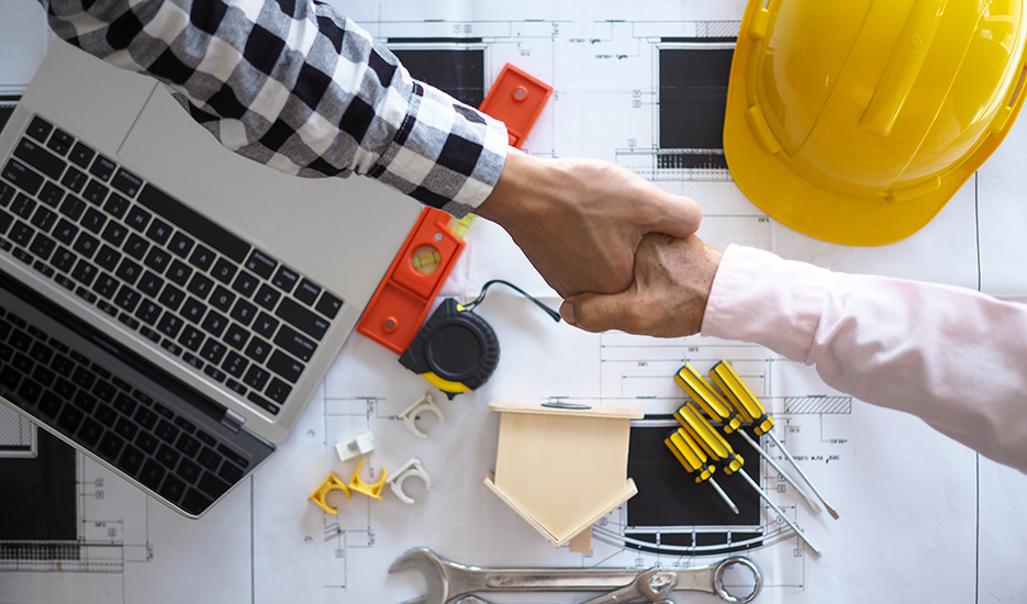 Two hands shaking over blueprints and general contractor tools