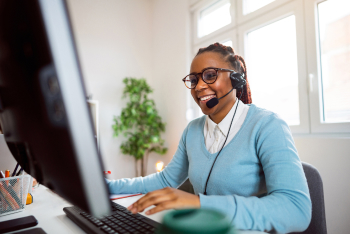 A woman with headphone and speakers working at her desk on her laptop smiling